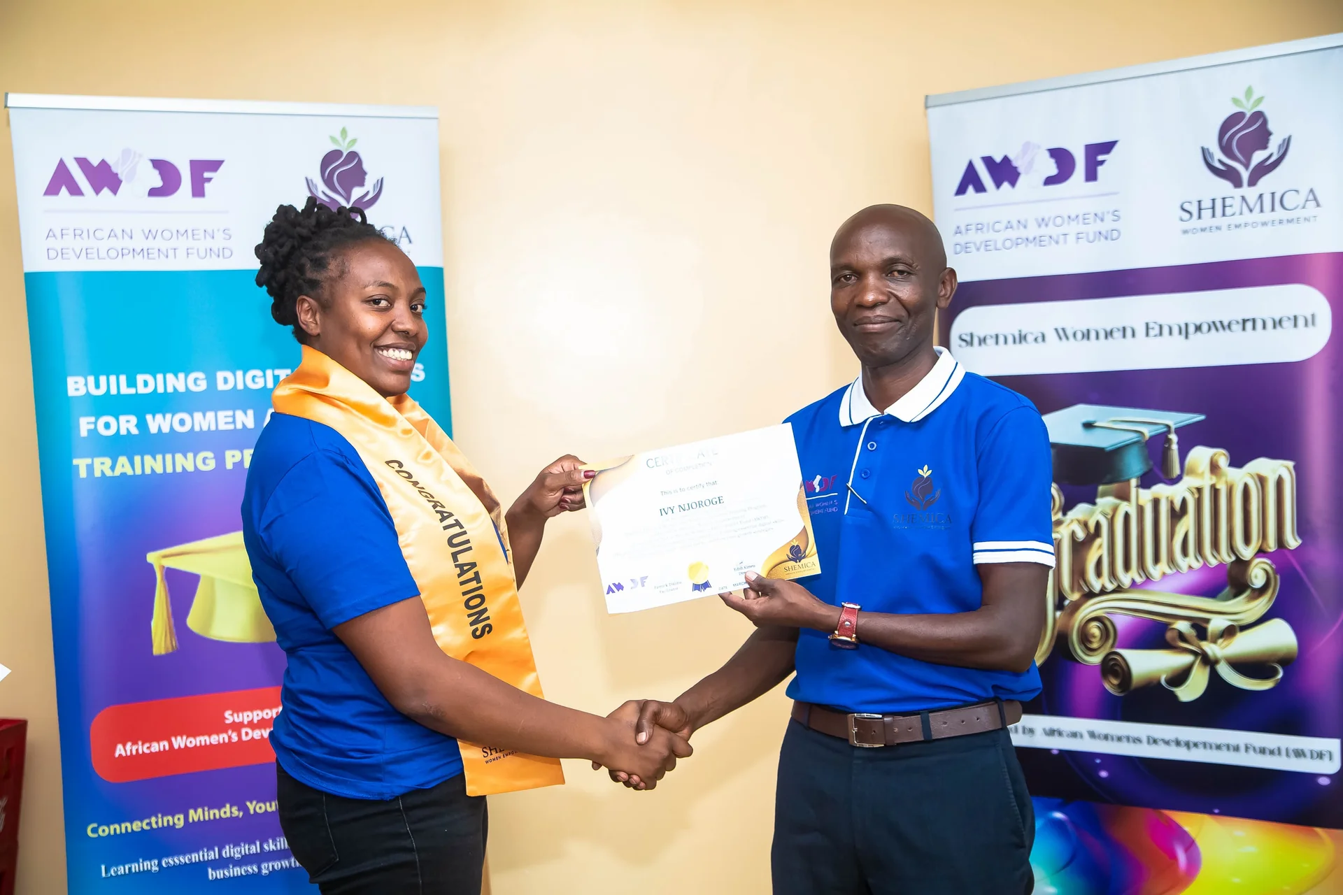 A smiling woman in a blue shirt and gold sash receives a certificate with a handshake.