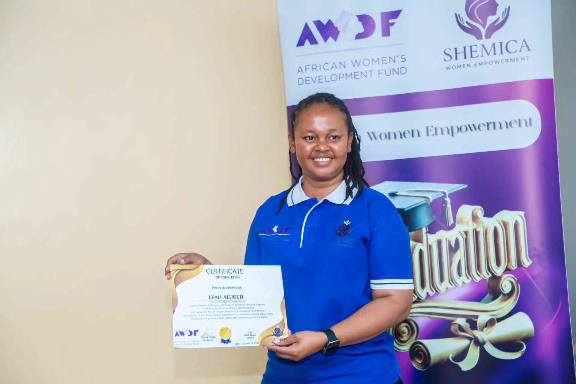 A woman in a blue Shemica shirt holds a certificate beside a graduation banner.
