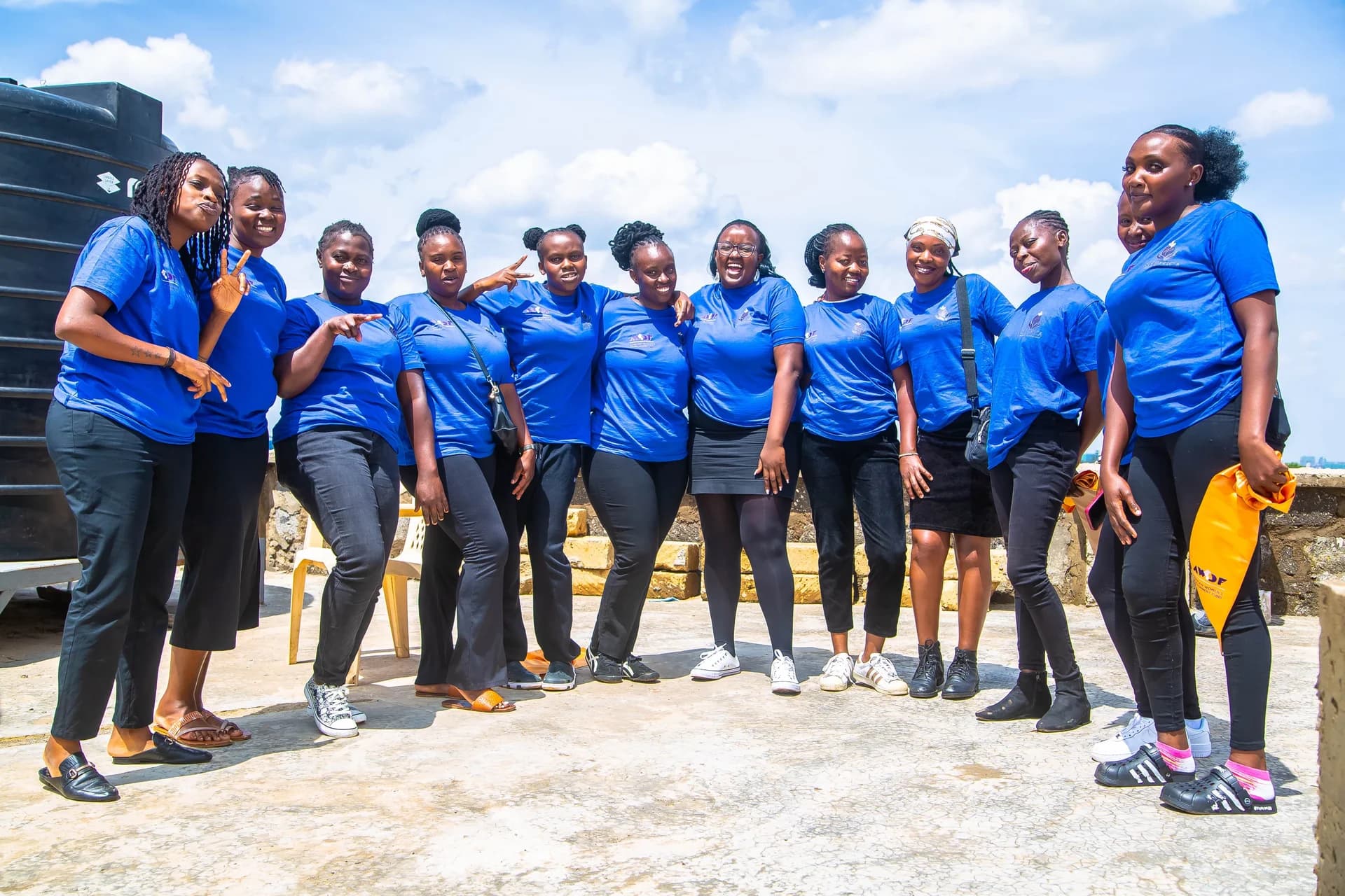 A group of women in blue Shemica shirts stands together outdoors, smiling and holding gold sashes.