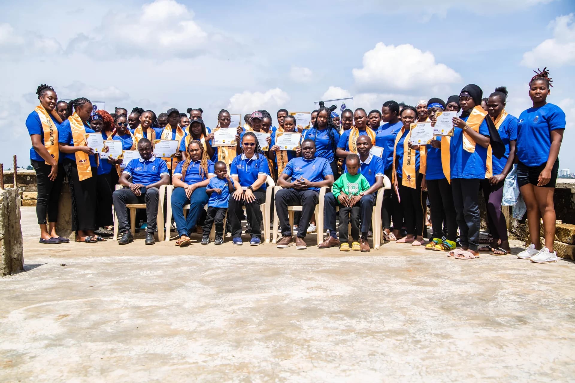Graduates in blue shirts and gold sashes gather outdoors with their certificates.